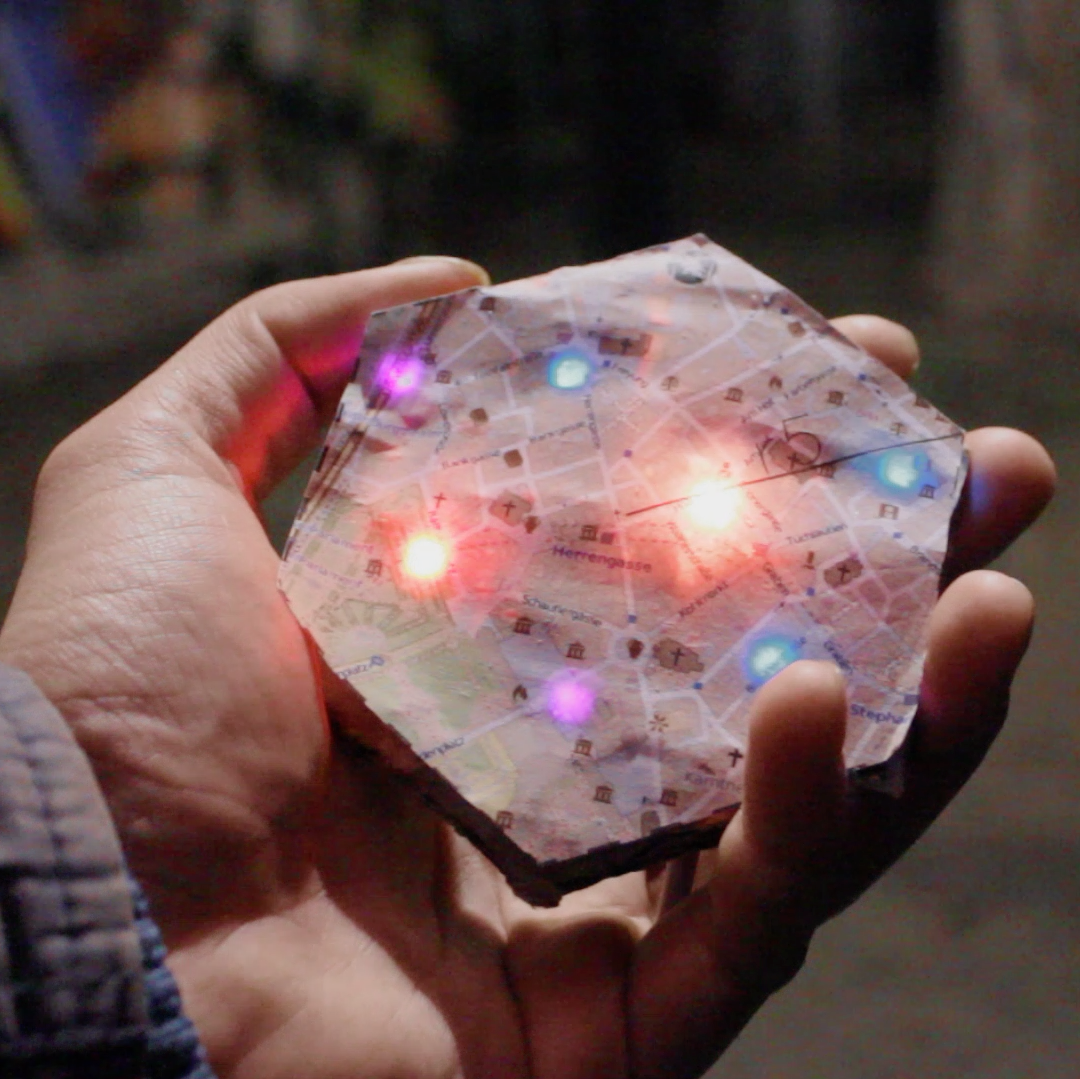 One hand holds a hexagonal game piece with a city map printed on it. Certain locations on the map are marked with different coloured lights. These are points of interest in the game. 