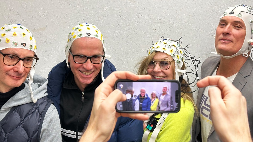 Some one else is taking a mobile phone picture of the Jet Lag All Stars and media artist Margarete Jahrmann are wearing EEG-hats. You can see both the screen of the phone and the real people behind it.
