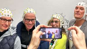 Some one else is taking a mobile phone picture of the Jet Lag All Stars and media artist Margarete Jahrmann are wearing EEG-hats. You can see both the screen of the phone and the real people behind it.