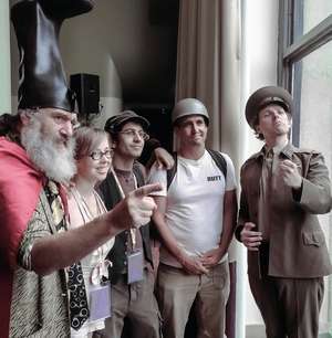 A group of people are standing in front of a window. Johannes Grentzfurthner is wearing a military uniform. Another person is wearing a welly on his head.
