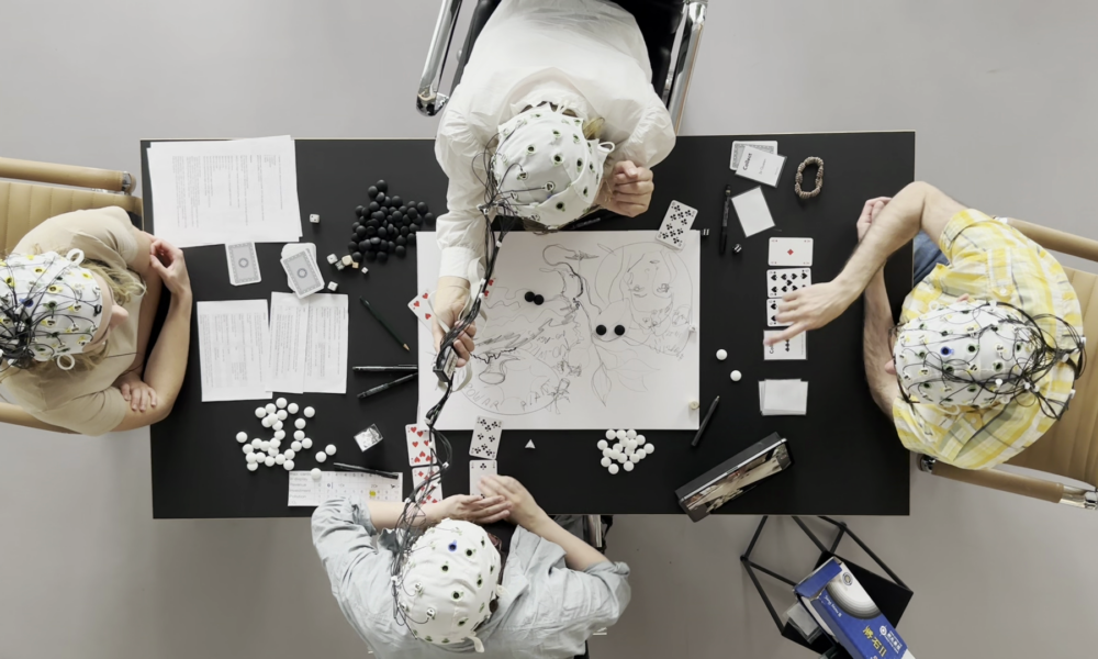 A picture of a table from above. There are four people sitting at the table, all wearing a EEG cap that is measuring their brain activites. On the table you can see the "Tree Game" prototype, consisting of a big piece of paper that two of the players are drawing on, some normal playing cards (Poker) and some black and white stones as well as some instructions for each player.