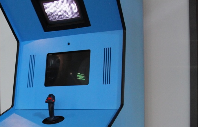 A picture of an exhibition space. There are people of all ages and genders playing different games on screens. They are sitting on tetris like blocks. You can read one of the game titles: "Chain Reaction". The vibes are chill and very colourful.