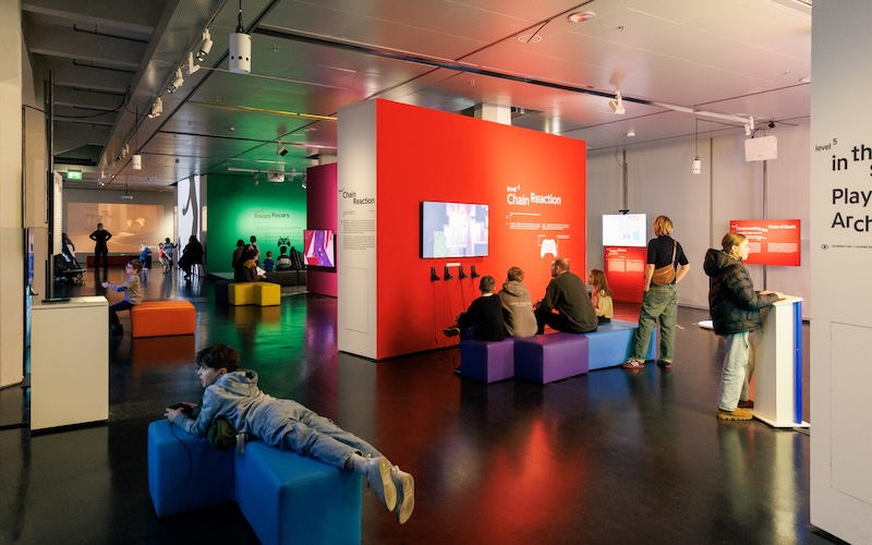 A picture of an exhibition space. There are people of all ages and genders playing different games on screens. They are sitting on tetris like blocks. You can read one of the game titles: "Chain Reaction". The vibes are chill and very colourful.