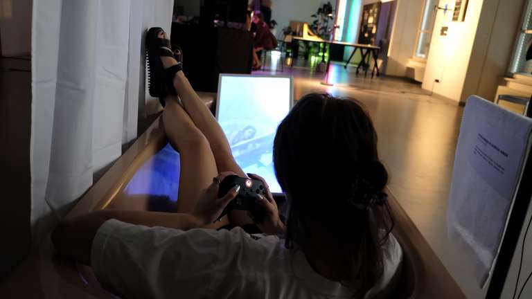 A person lying in a bathtub, holding a XBOX controller and looking at a screen on which you also see a person in a bathroom. The art style on the screen is very dreamy and soft.
